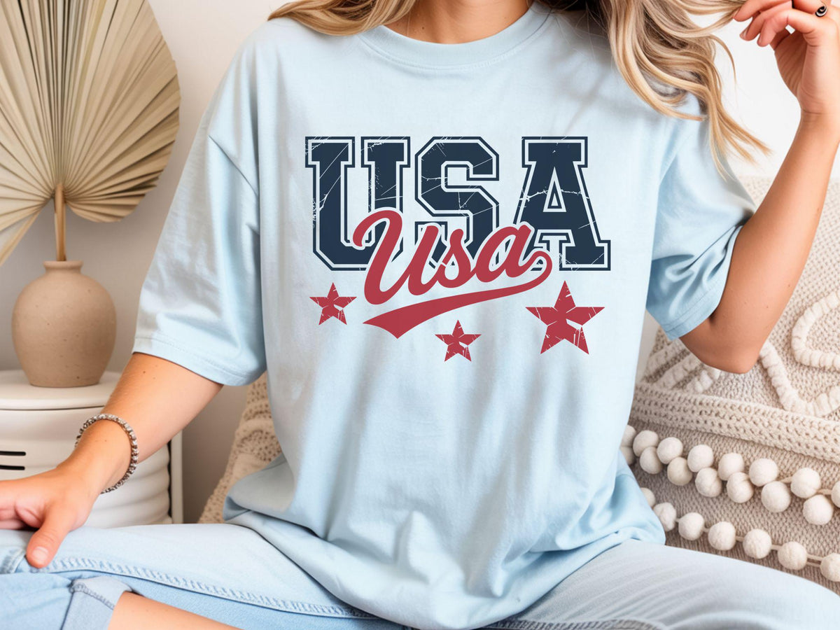May include: Light blue t-shirt with the word "USA" in navy blue and red, along with three red stars. The word "Usa" is in a cursive font. The shirt is a casual, patriotic design.