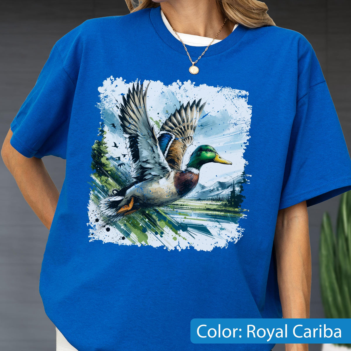 May include: Royal Cariba blue t-shirt featuring a detailed graphic of a flying duck with outstretched wings. The duck has green, brown, and white feathers, set against a backdrop of a landscape with mountains and trees. The shirt has a crew neck.