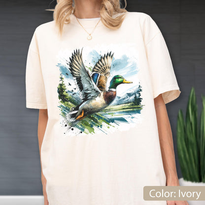 May include: Ivory-colored t-shirt featuring a watercolor illustration of a mallard duck in flight. The duck has green, blue, brown, and white feathers. The background includes a landscape with trees, water, and a blue sky. The text "Color: Ivory" is at the bottom.