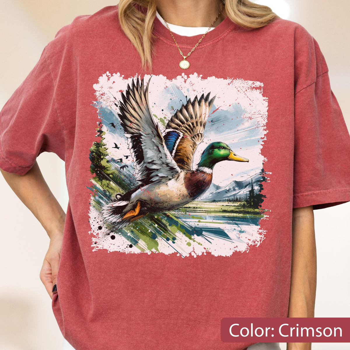 May include: Crimson-colored t-shirt featuring a vibrant graphic of a duck in flight. The duck has detailed feathers, a green head, and a yellow beak. The graphic is set against a landscape with mountains and water.