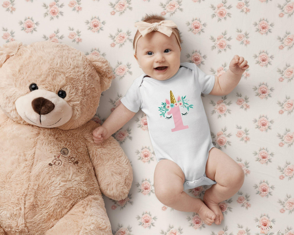 May include: A baby wearing a white onesie with a unicorn design and the number 1 in pink. The baby is lying on a floral patterned blanket next to a large tan teddy bear. A beige bow is in the baby's hair.
