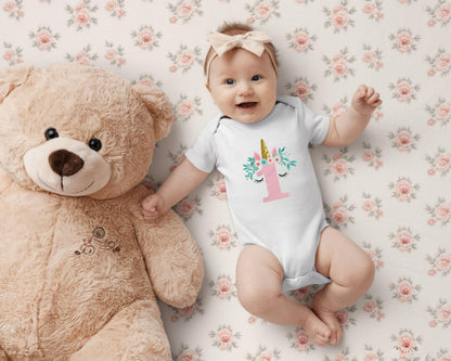 May include: A baby wearing a white onesie with a unicorn design and the number 1 in pink. The baby is lying on a floral patterned blanket next to a large tan teddy bear. A beige bow is in the baby's hair.