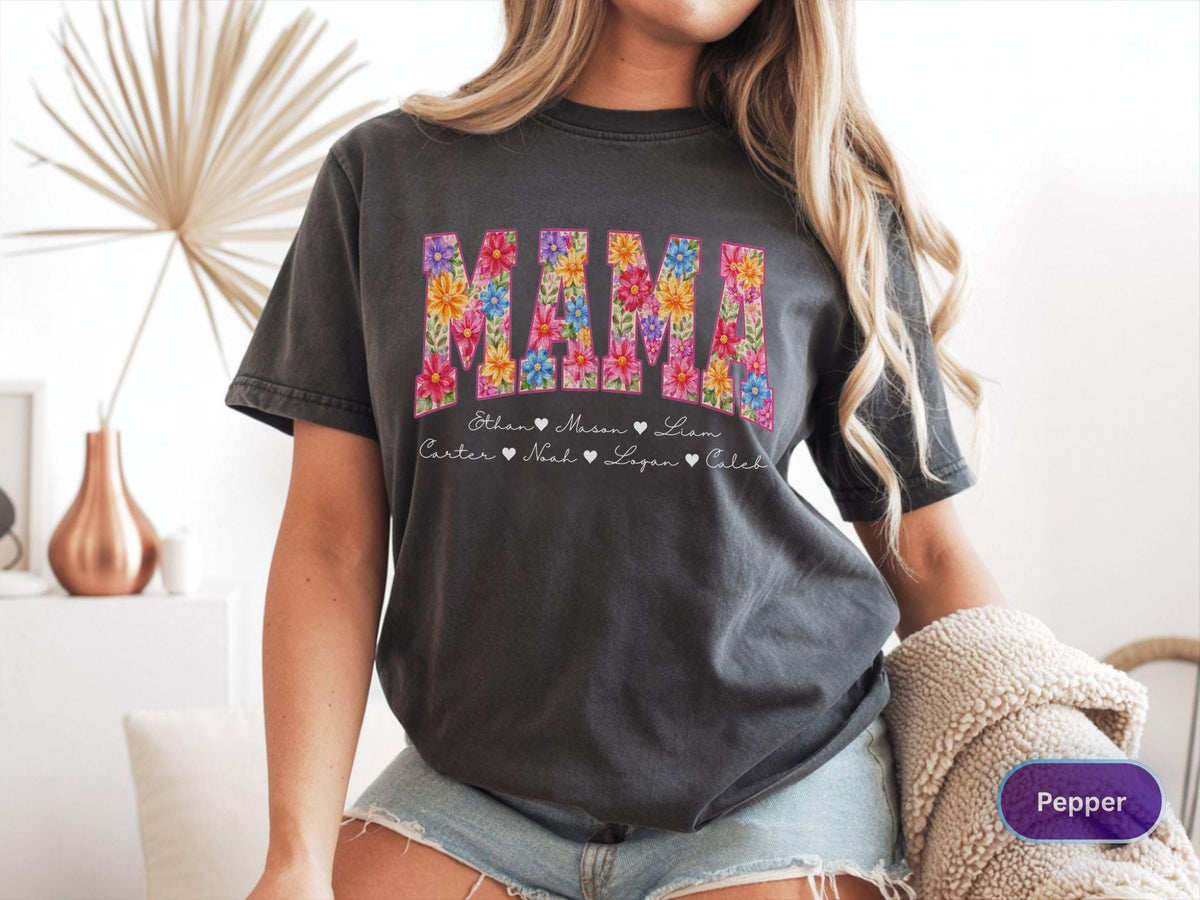 May include: Dark gray t-shirt with the word "MAMA" in colorful floral letters. Below the word "MAMA" are the names Ethan, Mason, Liam, Carter, Noah, Logan, and Caleb. The shirt is a casual style.