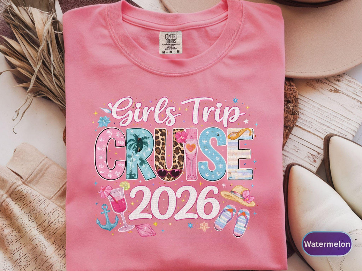 May include: A pink t-shirt with the words "Girls Trip Cruise 2026" in a colorful, decorative font. The design includes images of a palm tree, cocktail, anchor, and sandals. The shirt is a Comfort Colors brand.