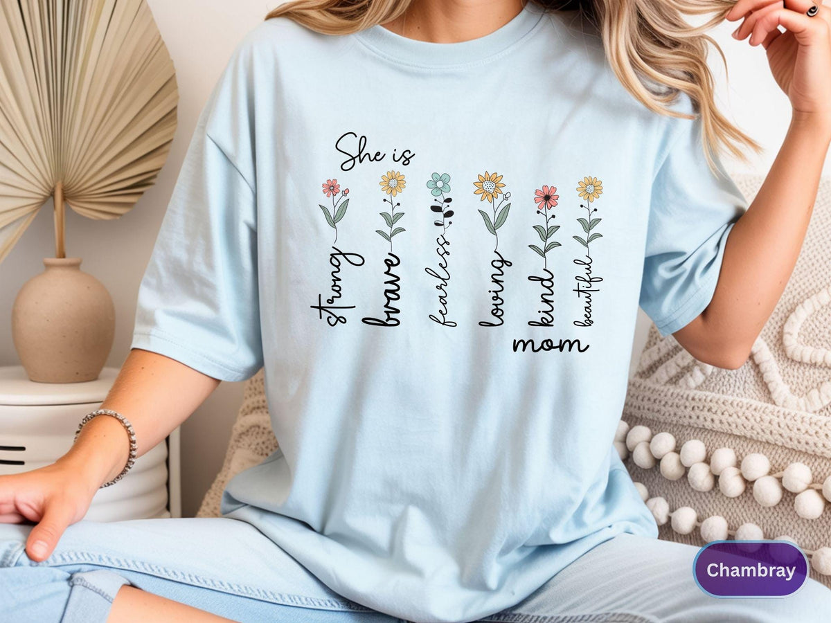 May include: Light blue t-shirt with the words "She is" above a row of floral stems. Each stem has a word: strong, brave, fearless, loving, kind, beautiful, and mom. The shirt is a soft, comfortable material.