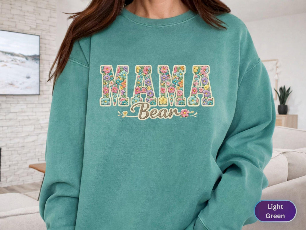 May include: A light green sweatshirt with the words "MAMA Bear" in a floral design. The letters are filled with colorful flowers, and the word "Bear" is in a cursive font. The sweatshirt has a crew neck and long sleeves.