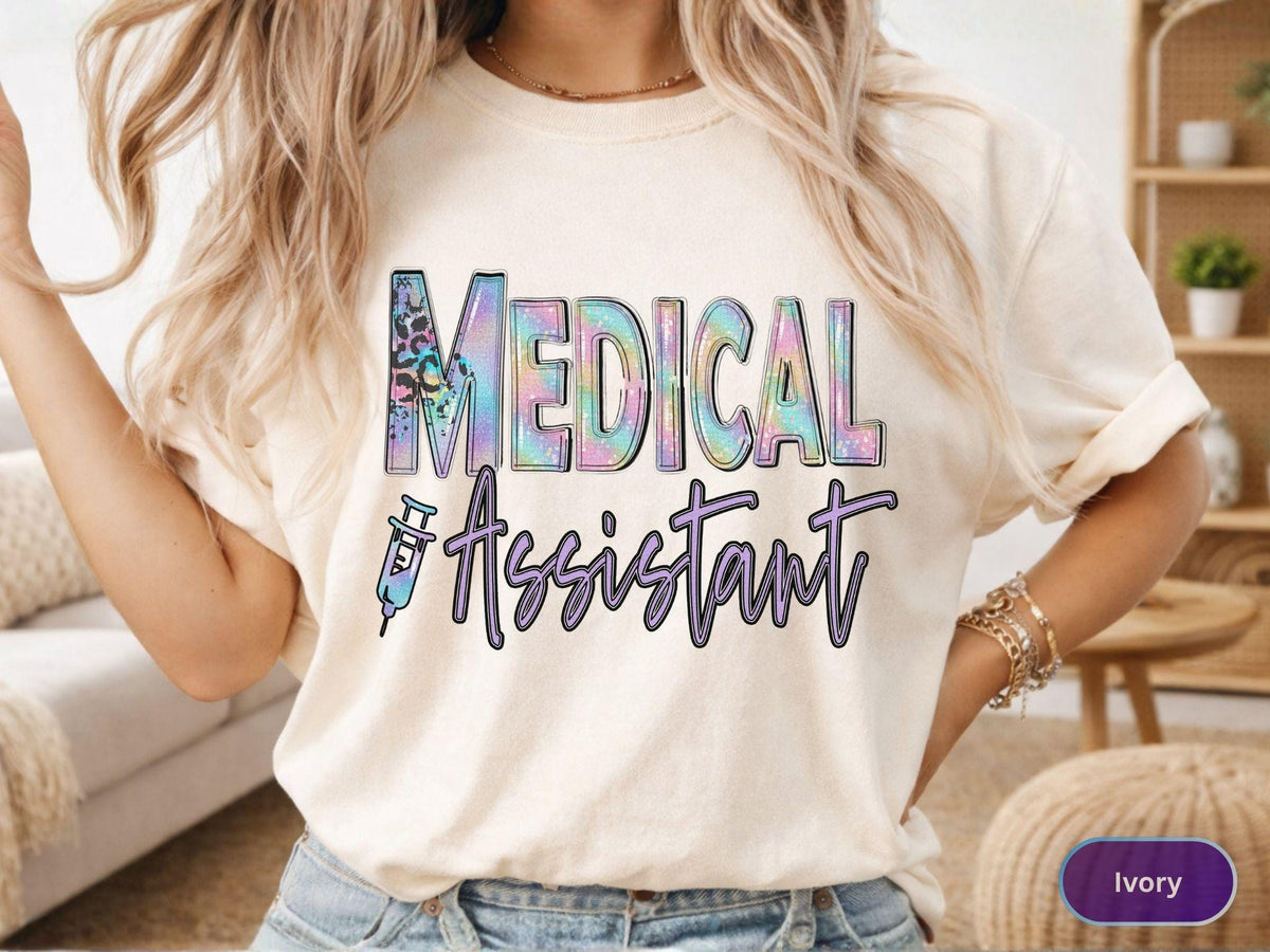 May include: Ivory-colored t-shirt with the words "MEDICAL ASSISTANT" in a colorful, holographic design. The word "MEDICAL" has a leopard print accent. A syringe graphic is included. The shirt is a casual style.