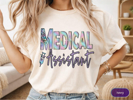 May include: Ivory-colored t-shirt with the words "MEDICAL ASSISTANT" in a colorful, holographic design. The word "MEDICAL" has a leopard print accent. A syringe graphic is included. The shirt is a casual style.