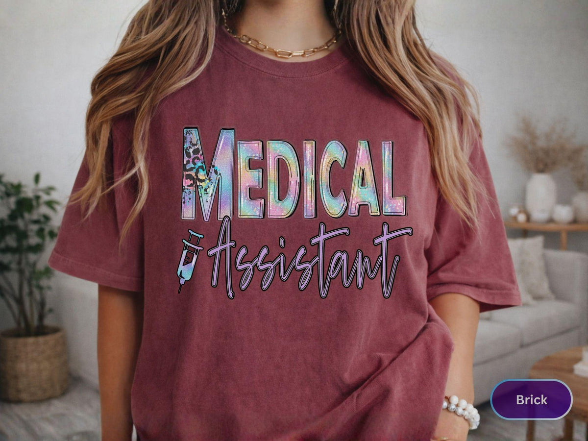 May include: Brick-colored t-shirt with the words "MEDICAL ASSISTANT" in a colorful, stylized font. The letter "M" has a leopard print design. A syringe graphic is below the text.