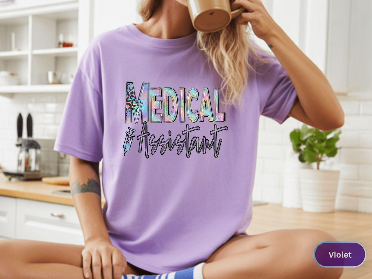 May include: A violet t-shirt with the words "MEDICAL ASSISTANT" in a colorful, holographic design. The shirt features a syringe graphic. The model is sitting down with a mug.