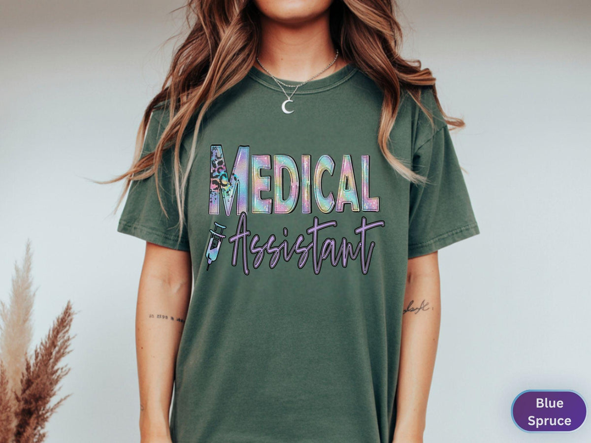May include: A spruce green t-shirt with the words "MEDICAL ASSISTANT" in a colorful, holographic design. The text is in a bold, stylized font. A syringe graphic is below the text. The shirt is a comfortable, casual style.