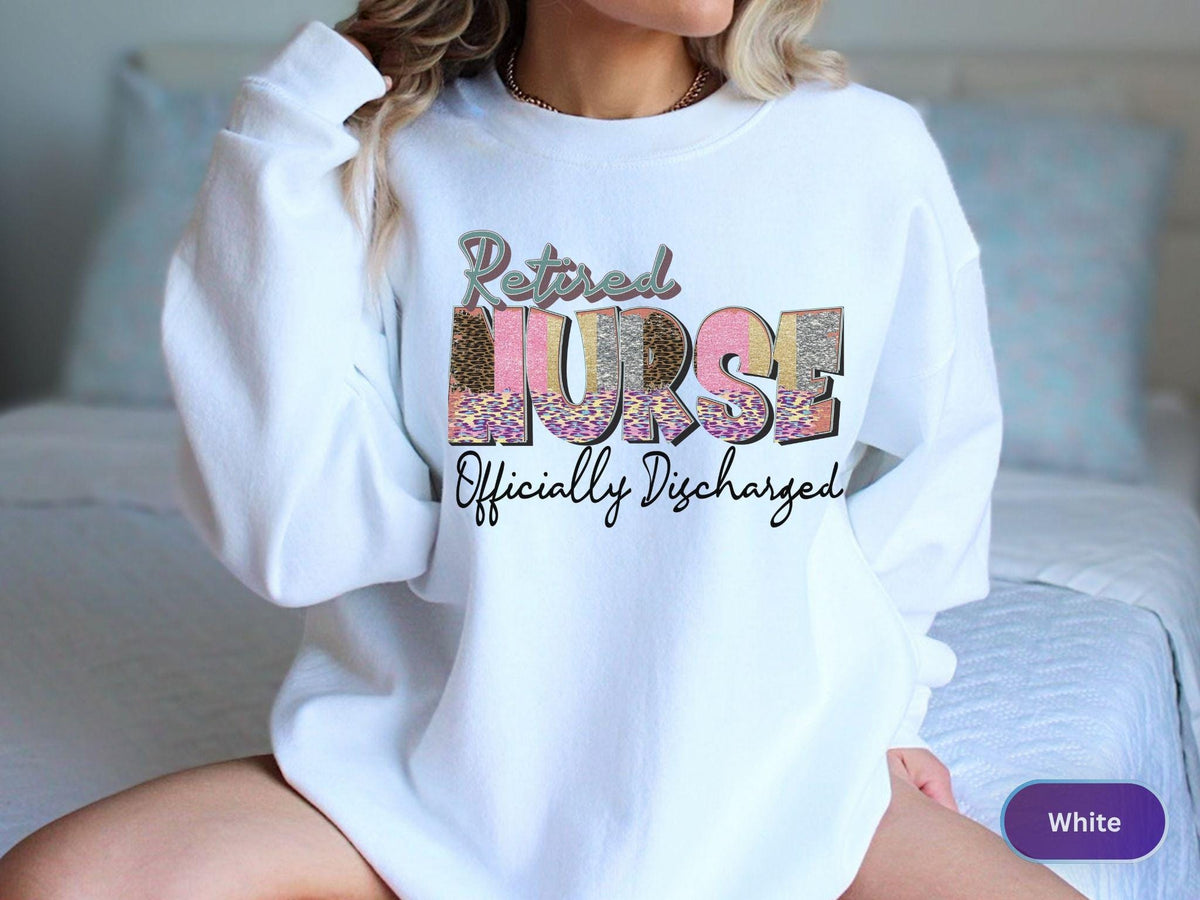 May include: White sweatshirt with the text "Retired Nurse Officially Discharged" in a mix of fonts and patterns. The word "NURSE" is in a leopard print and pink color. The sweatshirt is a casual style.