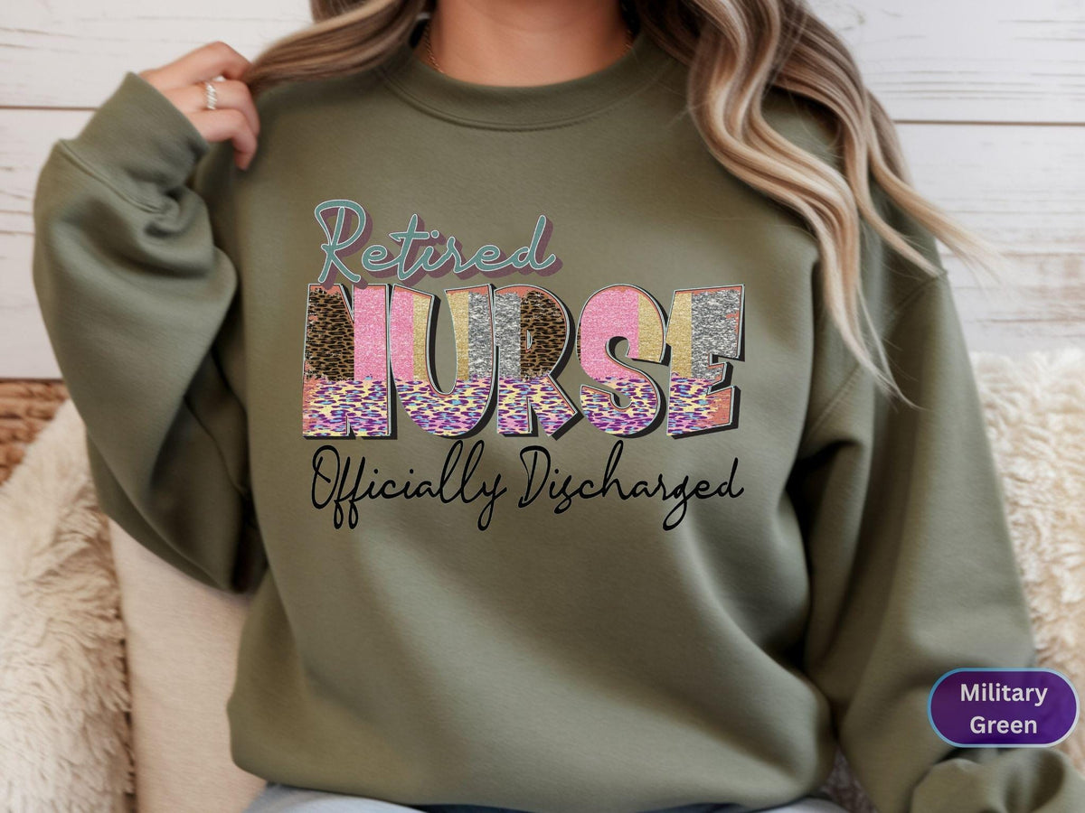 May include: Olive green sweatshirt with the text "Retired Nurse Officially Discharged" in a colorful, patterned design. The word "NURSE" is the focal point, with a mix of leopard print, pink, and glitter. The sweatshirt is a crew neck style.