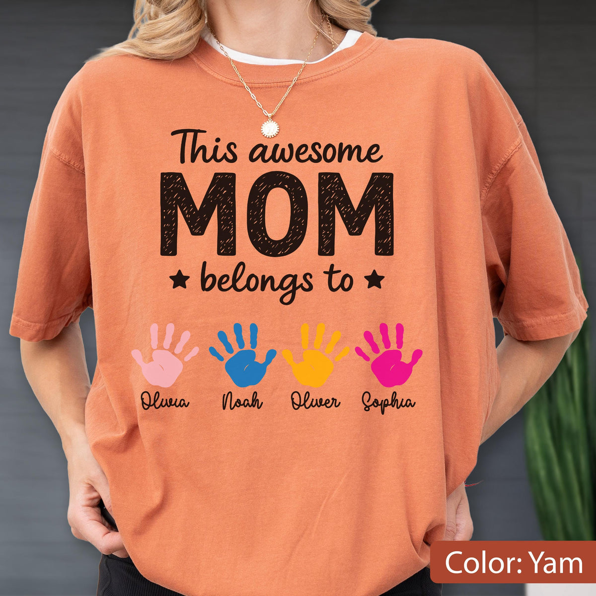 May include: A yam-colored t-shirt with the text "This awesome MOM belongs to" and four colorful handprints. Each handprint has a name below it: Olivia, Noah, Oliver, and Sophia. The shirt has a crew neck.