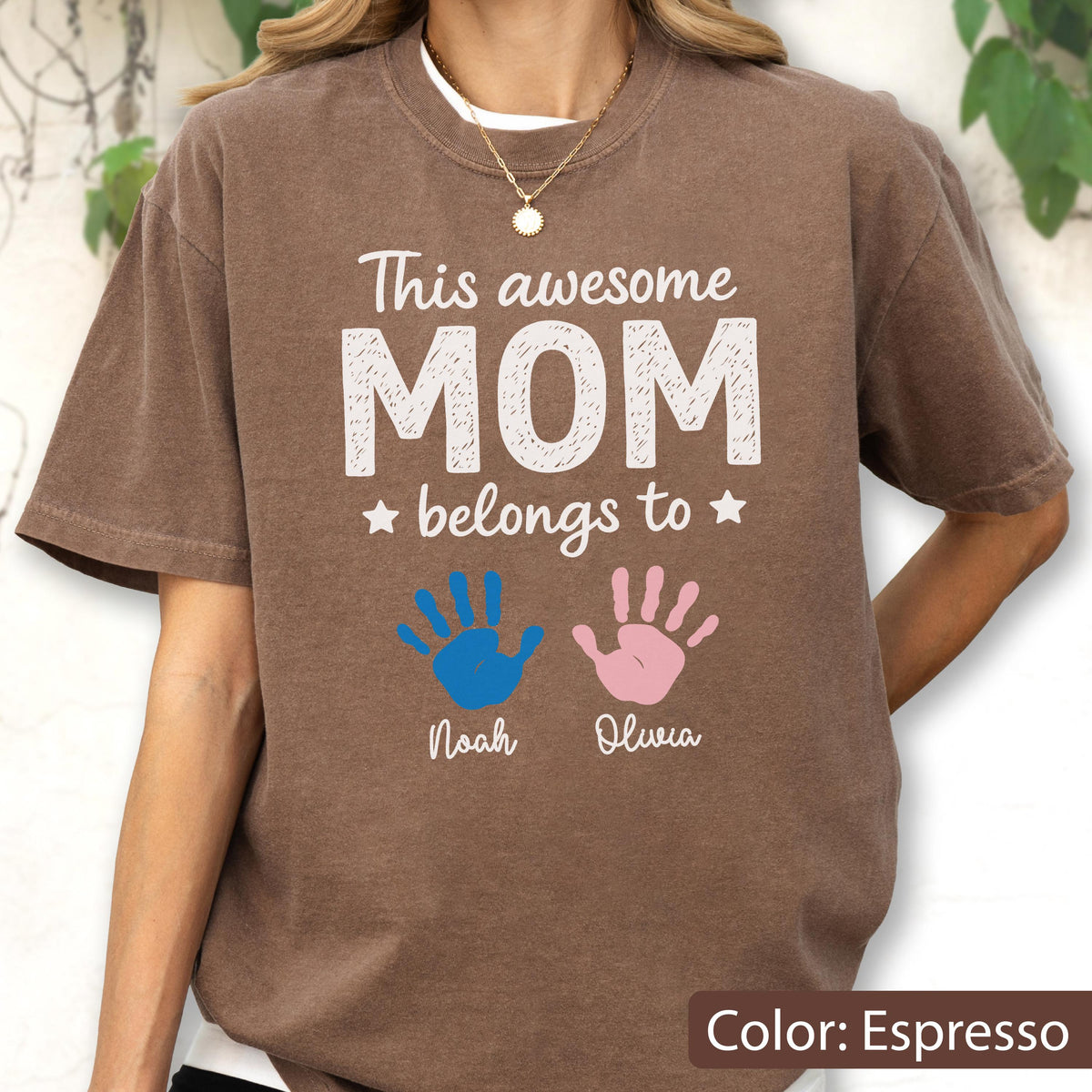 May include: Espresso-colored t-shirt with the text "This awesome MOM belongs to" above blue and pink handprints. Below the handprints are the names "Noah" and "Olivia". The shirt has a crew neck and short sleeves.