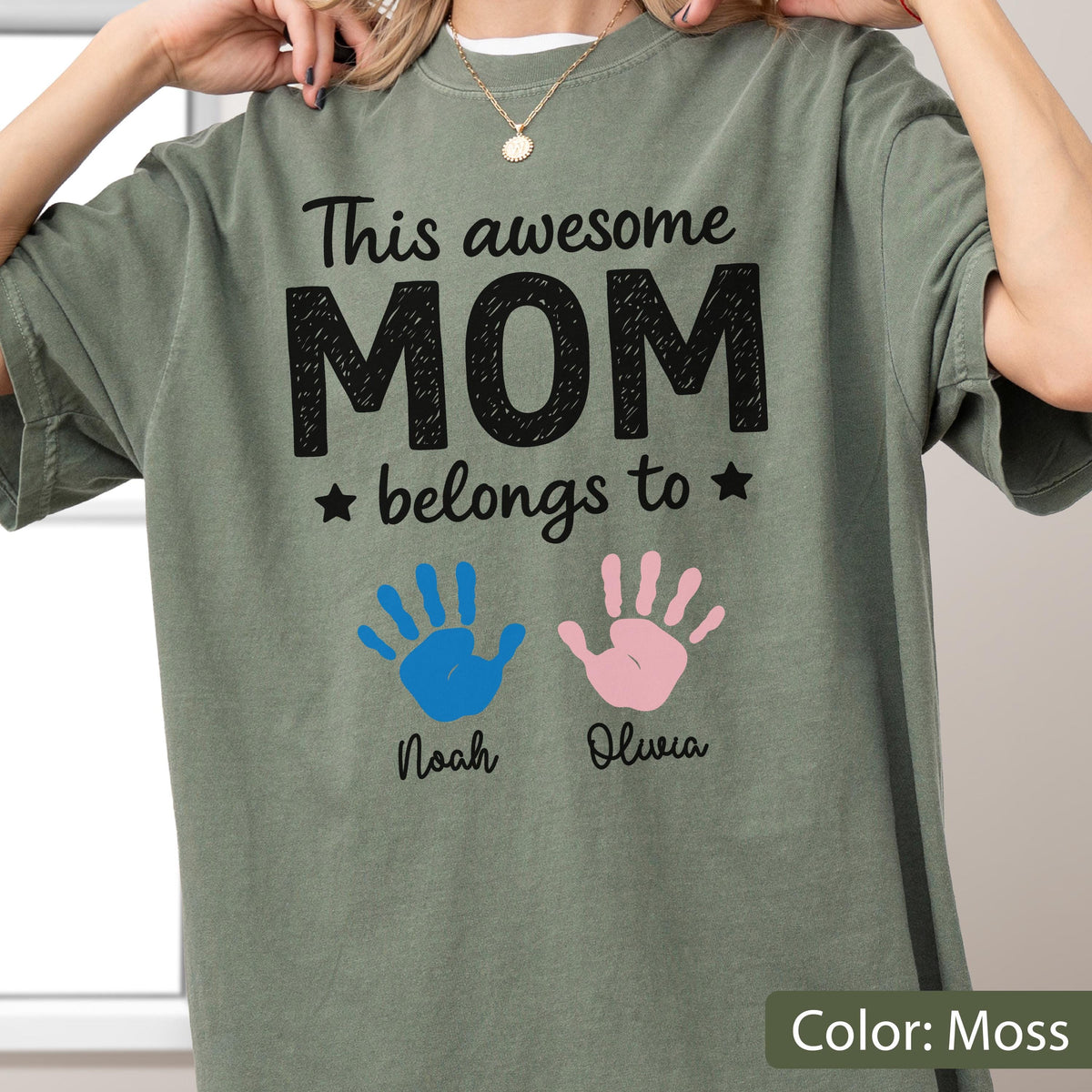 May include: Moss green t-shirt with the text "This awesome MOM belongs to" above blue and pink handprints. Below the handprints are the names "Noah" and "Olivia". The shirt is a solid color.