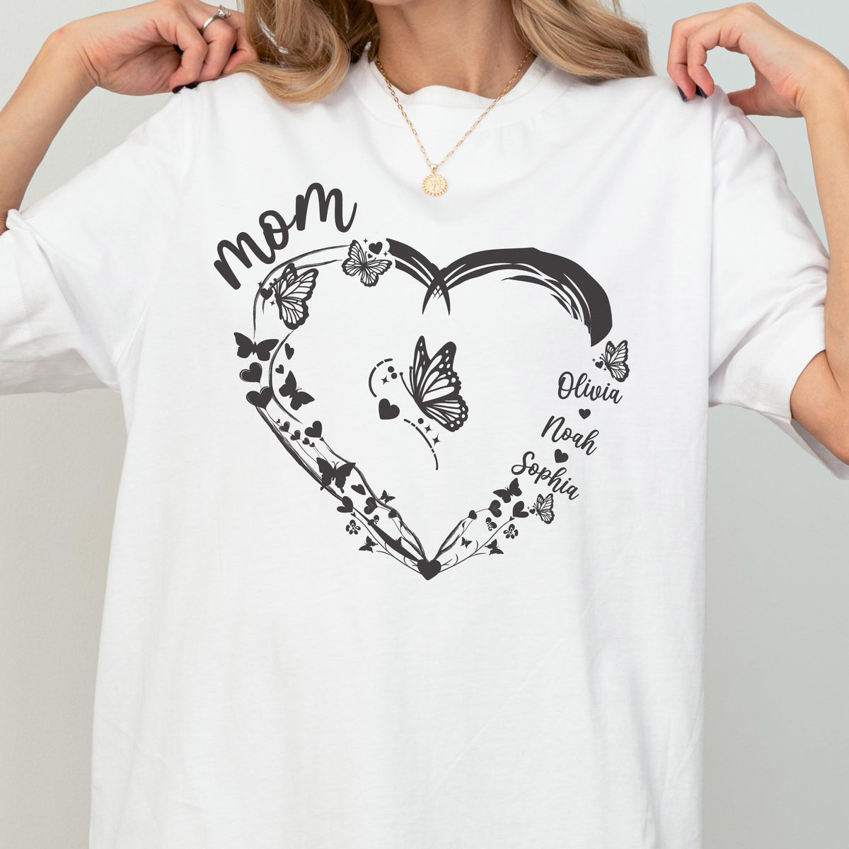 May include: White t-shirt featuring a black heart-shaped design with butterflies and the word "mom". The names "Olivia", "Noah", and "Sophia" are also included in the design. The shirt is a casual, everyday wear item.