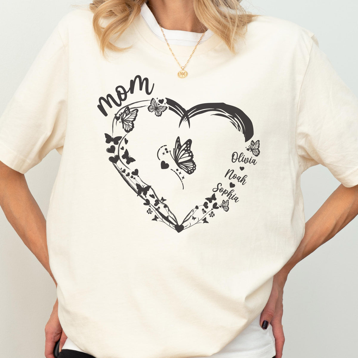 May include: Cream-colored t-shirt featuring a black heart design with butterflies and the word "mom". The names "Olivia", "Noah", and "Sophia" are printed on the right side of the heart. A gold necklace is visible.