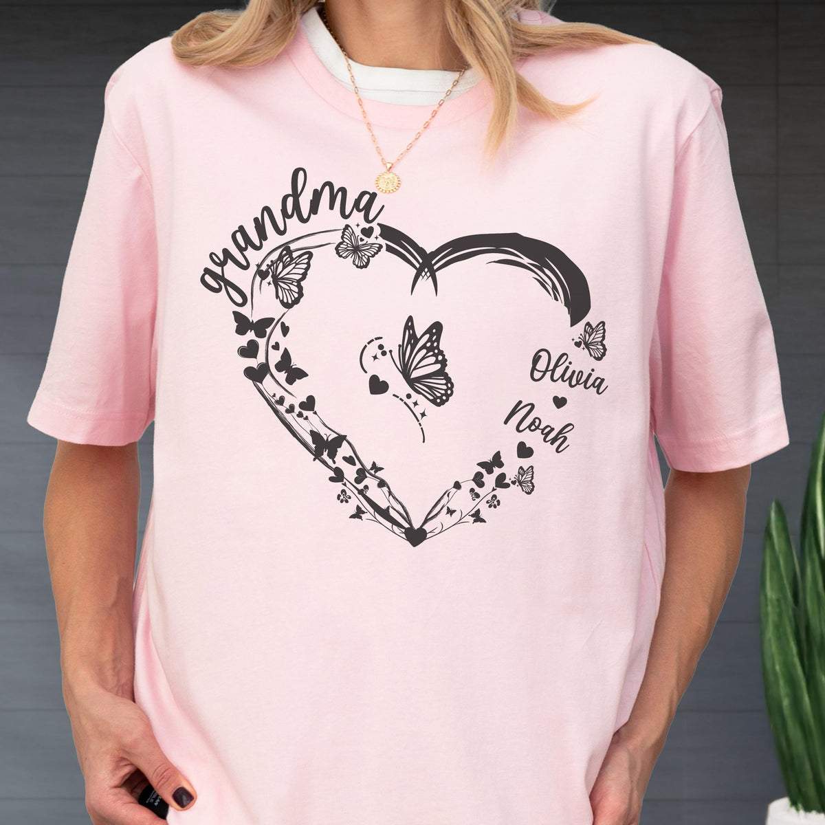 May include: A light pink t-shirt featuring a black heart-shaped design with butterflies and the word "grandma" in cursive. The names "Olivia" and "Noah" are also included. The shirt has short sleeves and a round neckline.