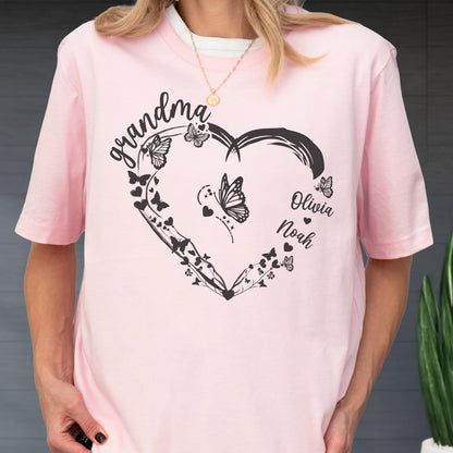 May include: A light pink t-shirt featuring a black heart-shaped design with butterflies and the word "grandma" in cursive. The names "Olivia" and "Noah" are also included. The shirt has short sleeves and a round neckline.