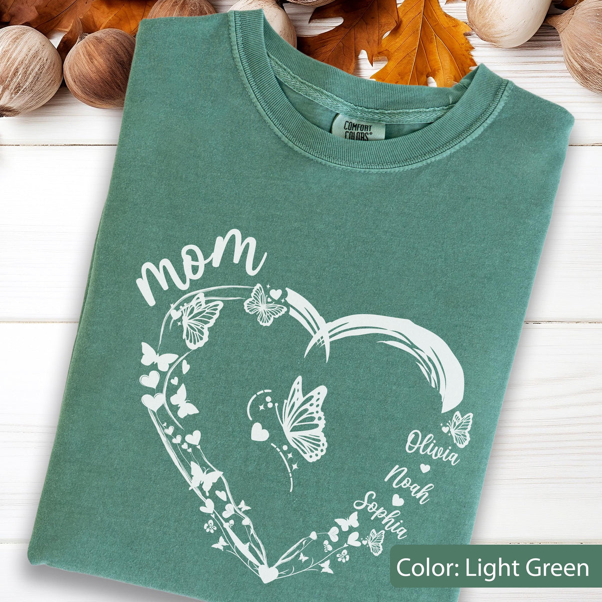 May include: Light green t-shirt with a white heart-shaped design filled with butterflies and the word "mom". The names Olivia, Noah, and Sophia are printed on the heart. The shirt is a Comfort Colors brand.