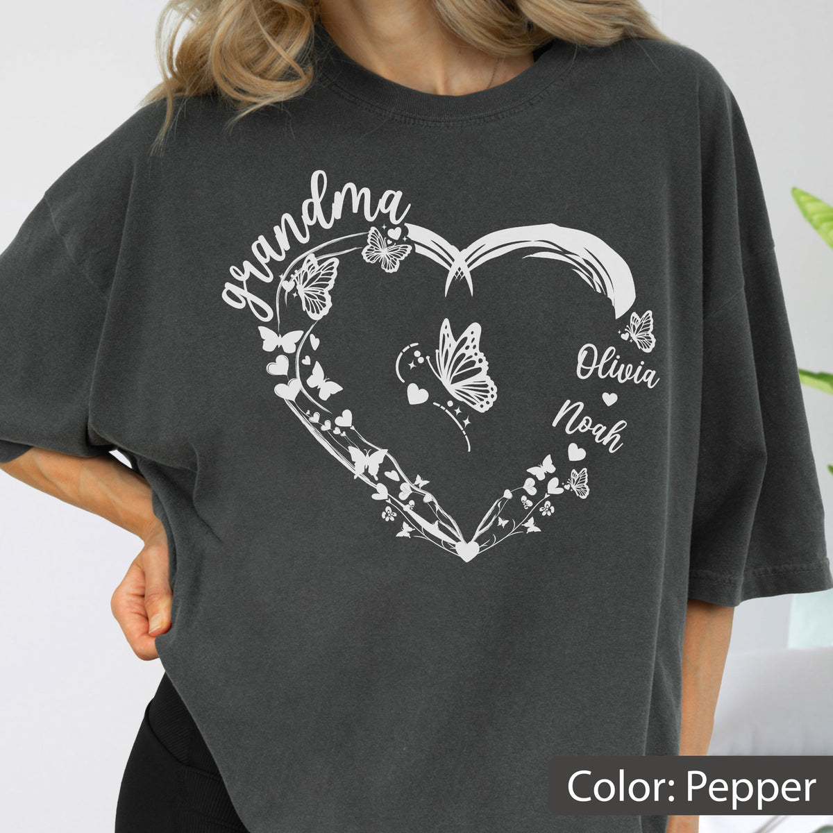 May include: Pepper-colored t-shirt featuring a white heart design with butterflies and the word "grandma" in script. The names "Olivia" and "Noah" are also included. The shirt is a relaxed fit.