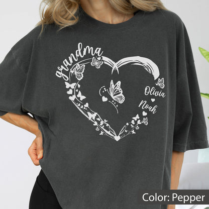 May include: Pepper-colored t-shirt featuring a white heart design with butterflies and the word "grandma" in script. The names "Olivia" and "Noah" are also included. The shirt is a relaxed fit.