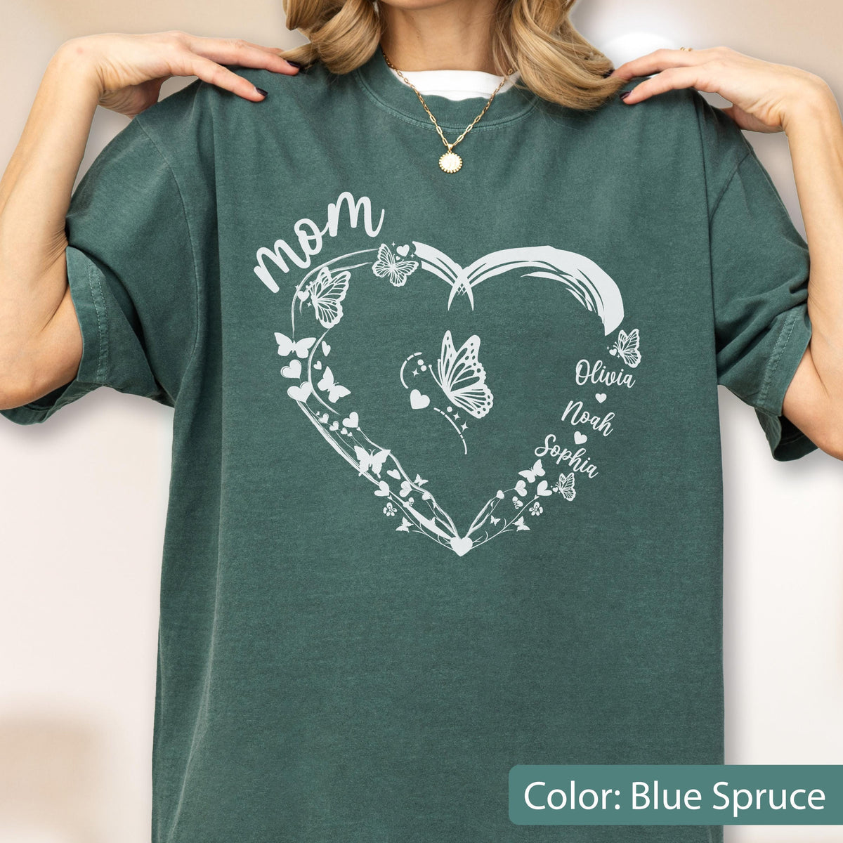 May include: Blue spruce-colored t-shirt with a white heart-shaped design. The design includes the word "mom" in script, butterflies, and the names "Olivia", "Noah", and "Sophia". The shirt is a solid color.