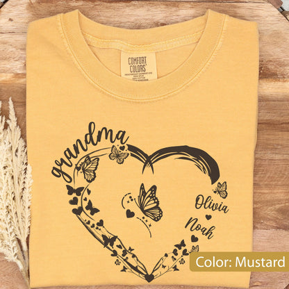 May include: Mustard-colored t-shirt with a heart-shaped design featuring butterflies and the word "grandma" in script. The names "Olivia" and "Noah" are also included. The shirt has a crew neck and a tag that says "Comfort Colors".