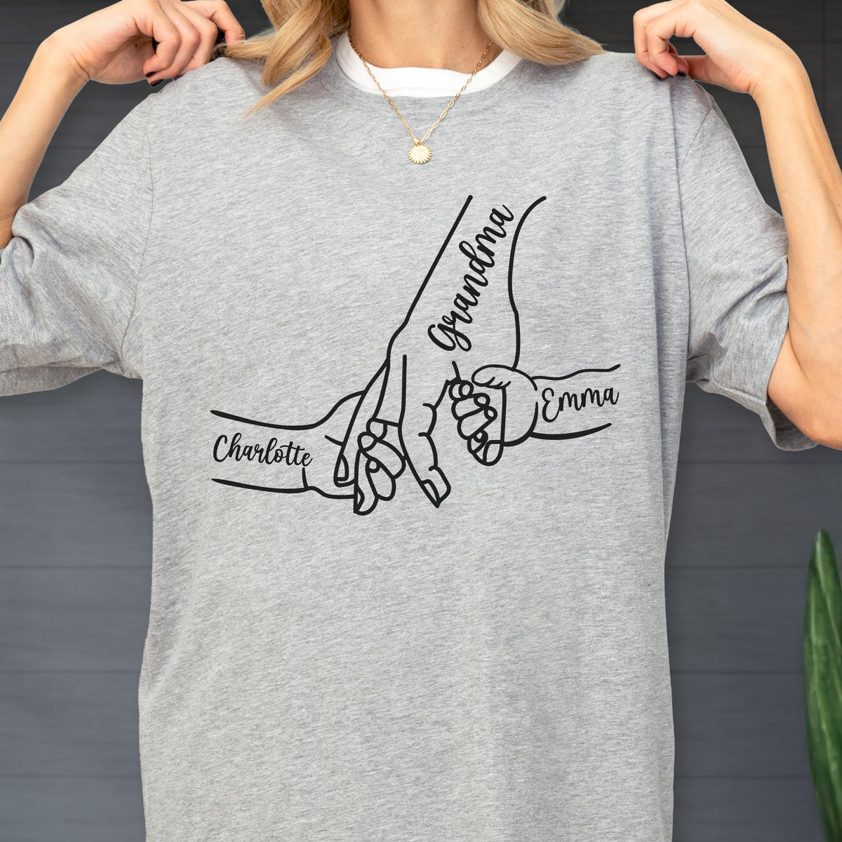 May include: A light gray t-shirt featuring a black graphic of hands holding each other. The word "Grandma" is written on one hand, with the names "Charlotte" and "Emma" on the other hands. The shirt is made of a soft material.