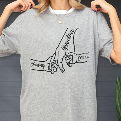 May include: A light gray t-shirt featuring a black graphic of hands holding each other. The word "Grandma" is written on one hand, with the names "Charlotte" and "Emma" on the other hands. The shirt is made of a soft material.
