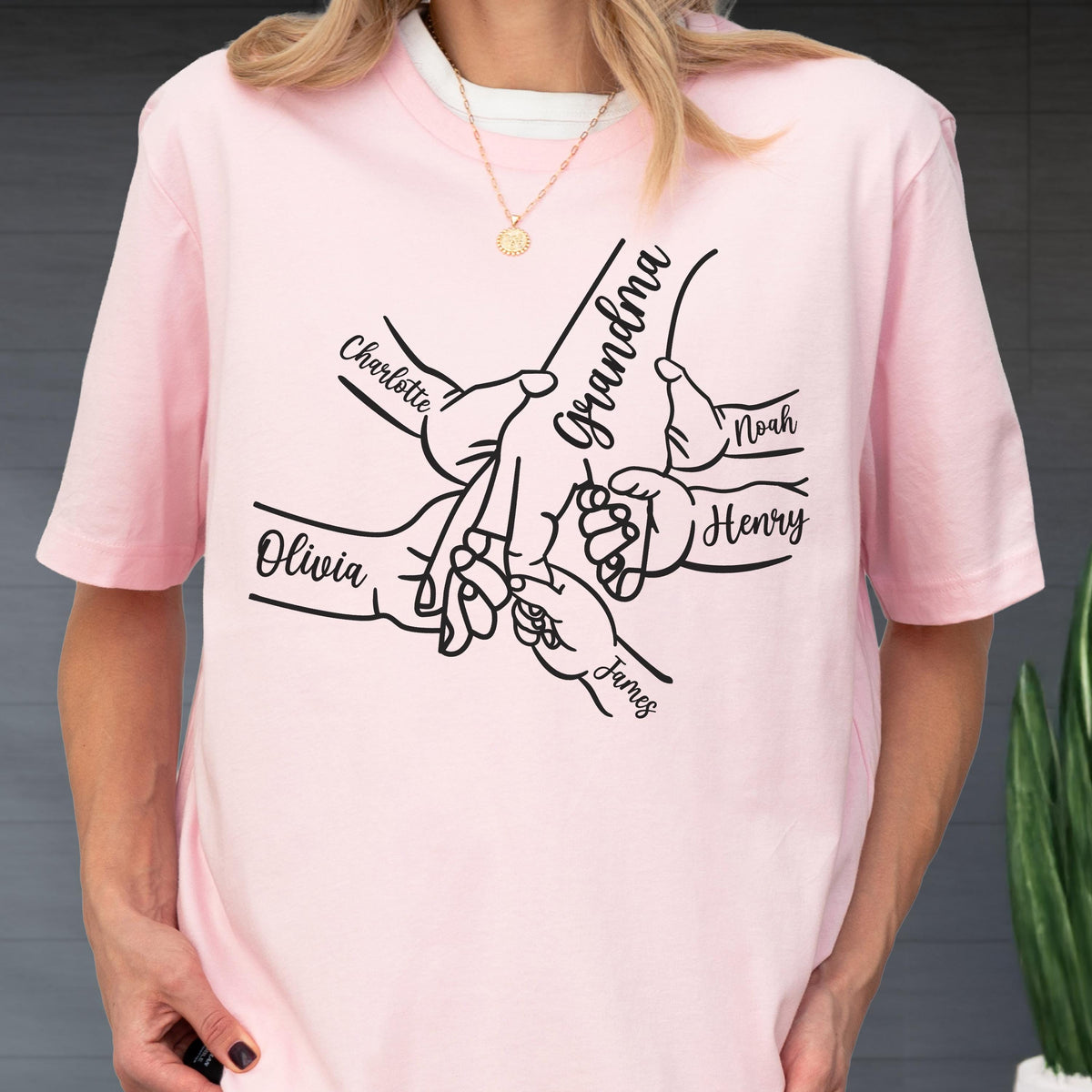 May include: A light pink t-shirt featuring a black graphic of hands intertwined, with the word "Grandma" and names "Charlotte", "Olivia", "Noah", "Henry", and "James" printed on it. The shirt is worn by a person.