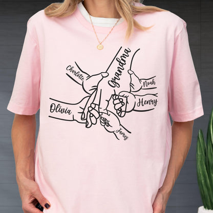 May include: A light pink t-shirt featuring a black graphic of hands intertwined, with the word "Grandma" and names "Charlotte", "Olivia", "Noah", "Henry", and "James" printed on it. The shirt is worn by a person.
