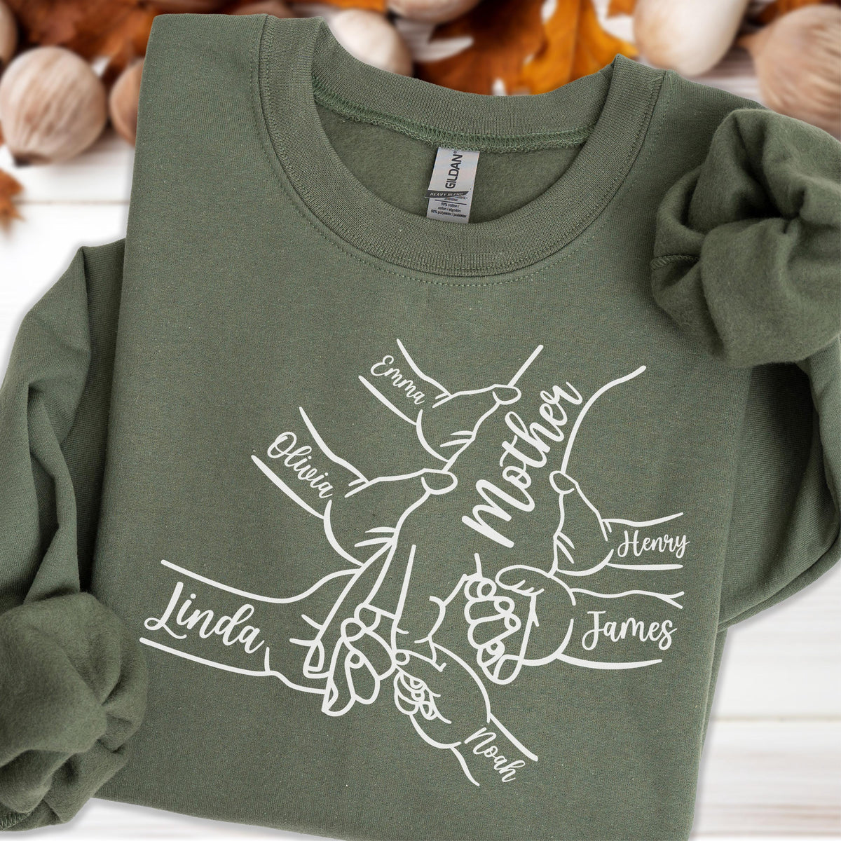 May include: An olive green sweatshirt with the word "Mother" in the center, surrounded by hands with names like "Emma", "Olivia", "Linda", "Henry", "James", and "Noah". The sweatshirt has a crew neck and long sleeves.