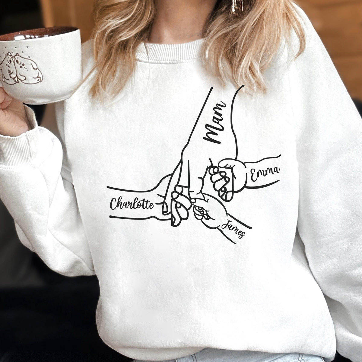 May include: White sweatshirt featuring a black graphic of hands holding each other, with the names "Mam", "Charlotte", "Emma", and "James" written on the hands. The person in the image is holding a white mug with a drawing of two dogs.