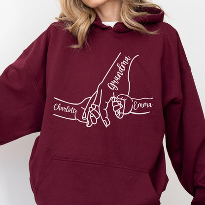 May include: Burgundy hoodie with a white graphic of hands holding each other. The word "Grandma" is written in cursive, with the names "Charlotte" and "Emma" below the hands. The hoodie has a drawstring hood and a front pocket.