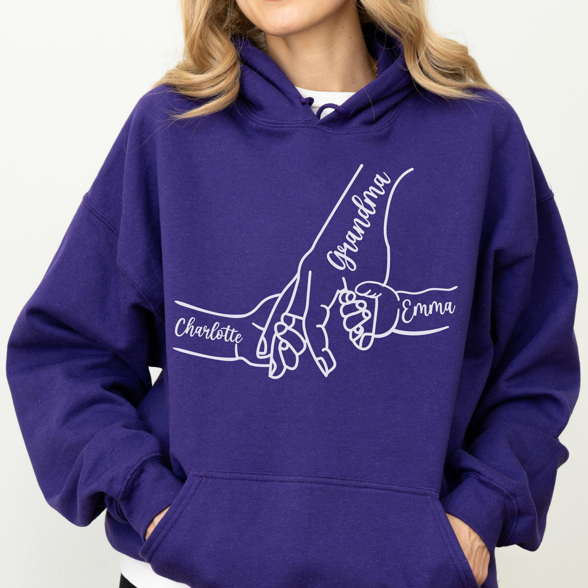 May include: A deep purple hoodie with a white graphic of three hands holding each other. The word "Grandma" is written on the largest hand, with the names "Charlotte" and "Emma" on the other hands. The hoodie has a hood and a front pocket.
