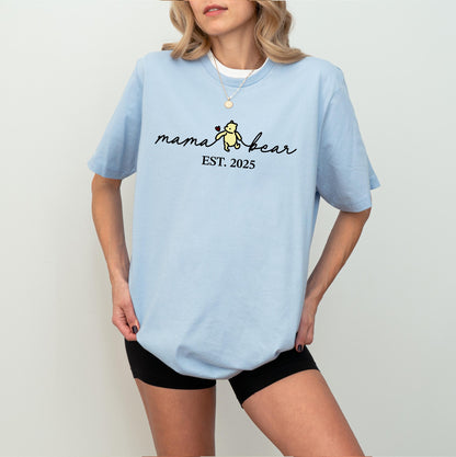 May include: A light blue t-shirt with the text "mama bear" in a script font, featuring a small bear graphic holding a heart. Below the text is "EST. 2025". The shirt is a short-sleeved, casual style.