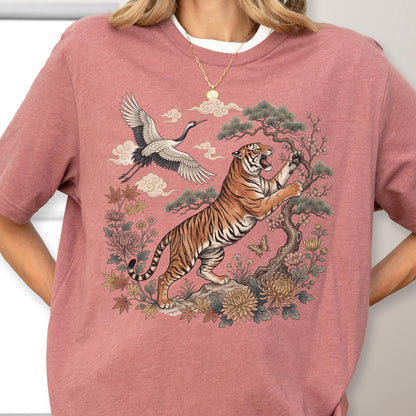 May include: A dusty rose t-shirt featuring a detailed illustration of a tiger, crane, and floral elements. The tiger is depicted with orange and black stripes, set against a backdrop of stylized clouds and foliage. The design has a vintage aesthetic.