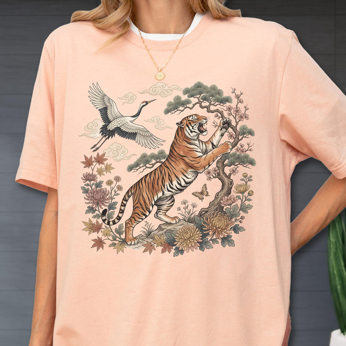 May include: Peach-colored t-shirt featuring a detailed illustration of a tiger climbing a tree, with a crane flying above. The design includes floral elements and a butterfly, creating a nature-inspired theme.