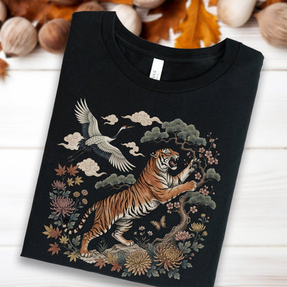 May include: Black t-shirt featuring a detailed illustration of a tiger, crane, and floral elements. The tiger is orange and black, with a fierce expression. The crane is white and gray, flying among clouds. The design includes trees, flowers, and butterflies.