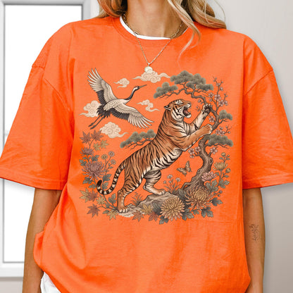 May include: Orange t-shirt featuring a detailed illustration of a tiger, crane, and floral elements. The tiger is depicted climbing a tree, with a crane flying above. The design includes clouds and a butterfly, creating a nature-inspired theme.