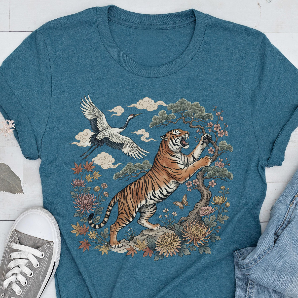 May include: Teal t-shirt featuring a detailed illustration of a tiger, crane, and floral elements. The tiger is orange and black, set against a backdrop of clouds, flowers, and a bonsai tree. A gray sneaker is visible in the lower left corner.