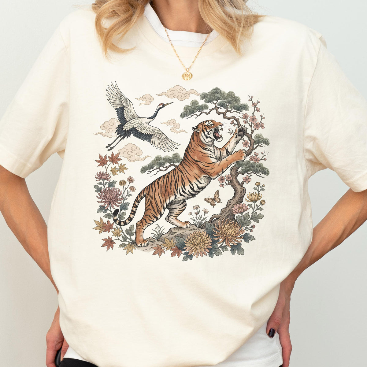 May include: Cream-colored t-shirt featuring a detailed illustration of a tiger, crane, and floral elements. The tiger is depicted with orange and black stripes, climbing a tree. The design includes a crane in flight and various flowers.