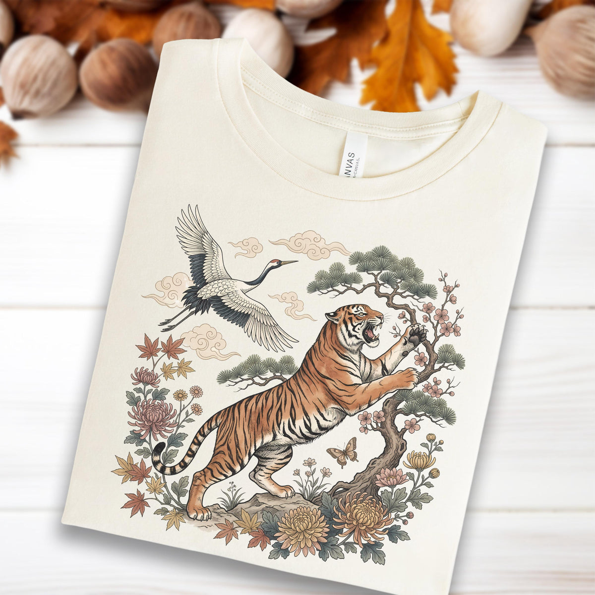 May include: Cream-colored t-shirt featuring a detailed illustration of a tiger, crane, and floral elements. The tiger is orange and black, reaching towards a flowering tree. A crane flies above, with clouds in the background. The design has an Asian-inspired aesthetic.