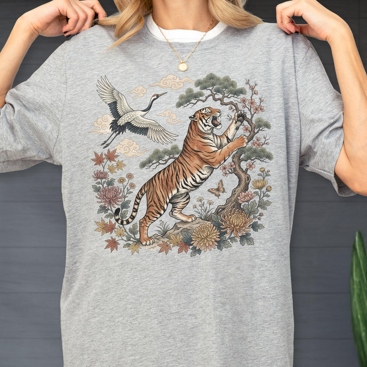 May include: A light gray sweatshirt featuring a detailed illustration of a tiger, crane, and floral elements. The tiger is depicted with orange and black stripes, reaching towards a tree. The design includes a crane in flight and various flowers.