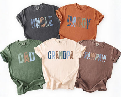 May include: Five short-sleeved t-shirts in various colors: charcoal, rust, olive green, cream, and brown. Each shirt features a different word in a distressed font: "UNCLE", "DADDY", "DAD", "GRANDPA", and "PAWPAW".
