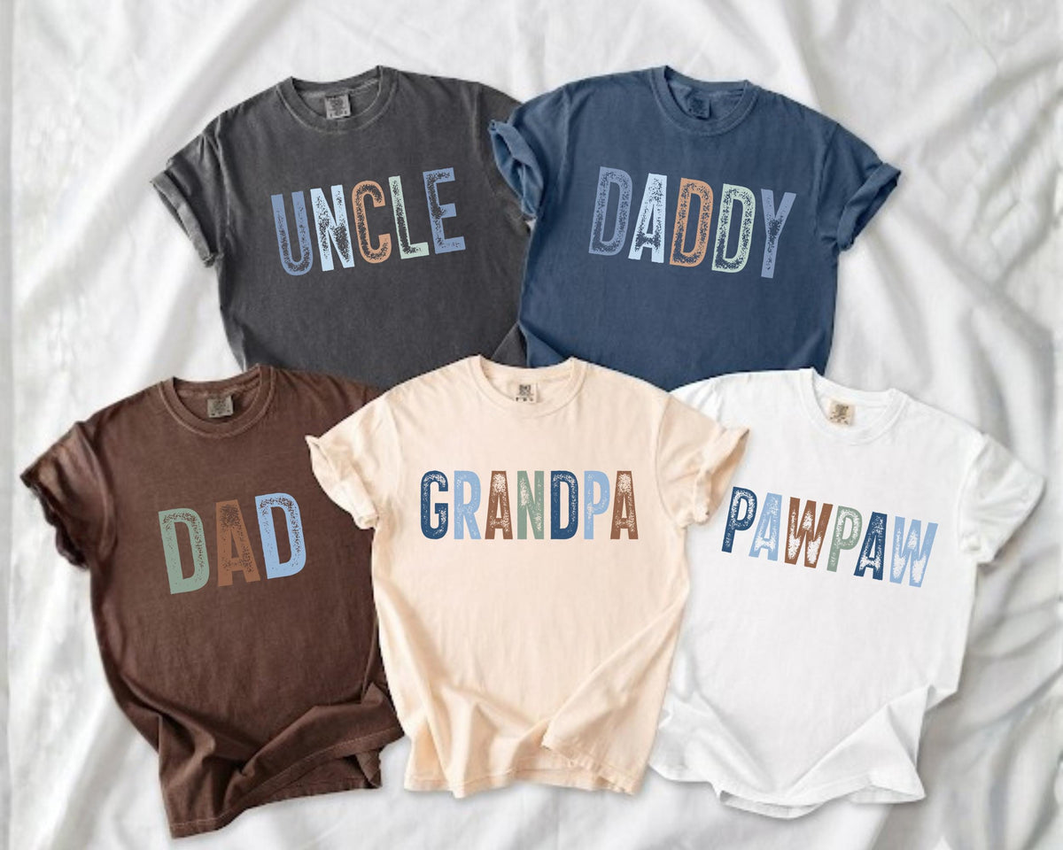 May include: Five short-sleeved t-shirts in various colors, including charcoal, navy, brown, cream, and white. Each shirt features a different word in a colorful, distressed font: "UNCLE", "DADDY", "DAD", "GRANDPA", and "PAWPAW".