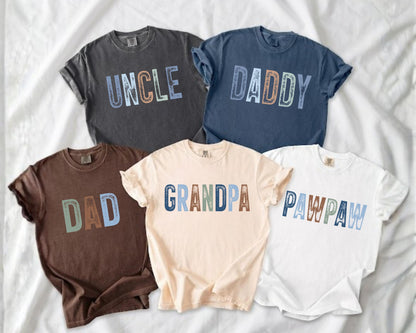 May include: Five short-sleeved t-shirts in various colors, including charcoal, navy, brown, cream, and white. Each shirt features a different word in a colorful, distressed font: "UNCLE", "DADDY", "DAD", "GRANDPA", and "PAWPAW".