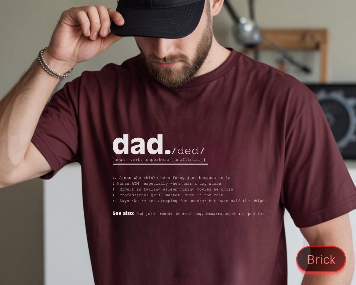 May include: A maroon t-shirt with the word "dad" in white, followed by a phonetic spelling and a definition. The definition includes humorous descriptions. The shirt is a crew neck style.